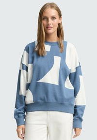 Jonge vrouw in een blauw-wit abstract patroon sweatshirt en witte broek, staand tegen een effen lichtgrijze achtergrond.