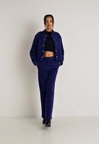 Ναυτικό μπλε καμπαρντίνα με κουμπιά, σε συνδυασμό με ταιριαστά παντελόνια καμπαρντίνας. Το σύνολο διαθέτει ένα κοντό μαύρο τοπ από κάτω.