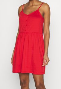 Robe sans manches rouge en coton. Elle dispose d'un corsage ajusté avec des boutons et d'une jupe évasée avec une taille froncée.