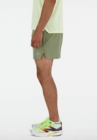 Hellgrüne Sportshorts mit seitlichem Schlitz und hellgrauem Logo, kombiniert mit neon-gelben und schwarzen Laufschuhen. Schlicher Hintergrund.