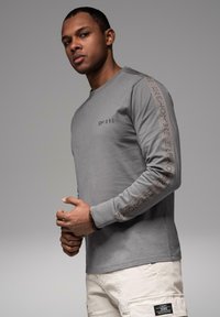 Muž v šedé dlouhém rukávu s textem "OMBRE" na hrudi a rukávech, béžových cargo kalhotách a s korálkovým náramkem, pózující na šedém pozadí.