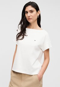 Λευκό κοντομάνικο τοπ με λαιμόκοψη βάρκα και κεντητό λογότυπο σε κόκκινο, λευκό και μπλε. Το ύφασμα φαίνεται λείο με άνετη εφαρμογή.