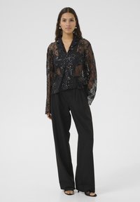 Bluză neagră cu paiete, cu guler și mâneci lungi din material transparent, asortată cu pantaloni negri largi și pantofi negri cu vârful deschis.