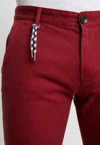 Rode textuurbroek met knoopsluiting en een decoratieve trekkoord met blauw, wit en rode patronen. Close-up van de taille.