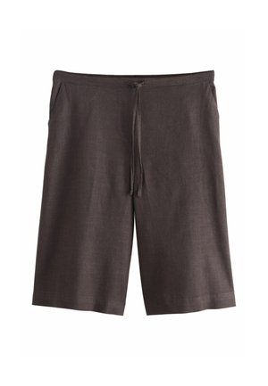Vrouw met gevlochten knotkapsel, draagt een mouwloos wit topje en losse donkere knielange korte broek, staand met handen in de zakken.
