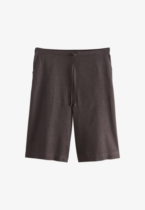 Kvinde med flettet knold, iført hvid ærmeløs top og løse mørke knælange shorts, står med hænderne i lommerne.