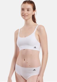Soutien-gorge de sport blanc avec des fines bretelles, texture lisse, et un logo Adidas noir sur le côté. Assorti à un sous-vêtement blanc.