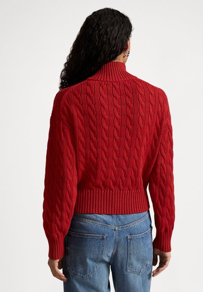 Γυναίκα με μακριά σγουρά μαλλιά, φορώντας κόκκινο πλεκτό πουλόβερ με κόμπους και μπλε τζιν, που φαίνεται από πίσω πάνω σε απλό φόντο.