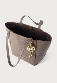 Rjava strukturirana torba tote z teksturirano zunanjostjo, dvema ročajema in zlatim obeskom z logotipom. Notranjost je temna z minimalnimi žepi.