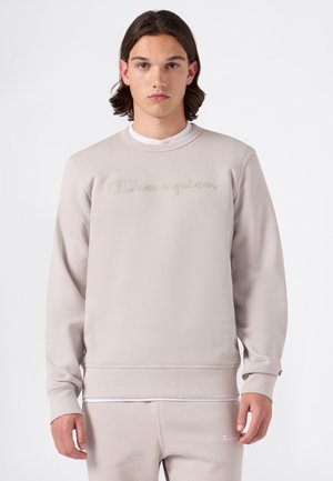 Giovane uomo con capelli di media lunghezza che indossa una felpa Champion beige chiaro e pantaloni della tuta coordinati, in piedi davanti a uno sfondo bianco.