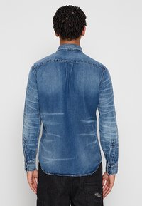 Homme aux cheveux courts et torsadés, portant une chemise en denim bleu délavé à manches longues et un pantalon noir, vu de dos.