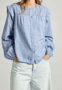 Hellblaues Hemd mit Knopfleiste, Puffärmeln, Spitzenbesatz und einem Einsatz mit vertikalen Falten, gefertigt aus weichem Baumwollstoff.