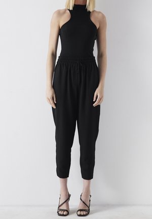 Mujer con top negro sin mangas, pantalones cortos negros anchos con cintura ajustable y sandalias negras de tacón alto con tiras, de pie junto a una pared blanca.