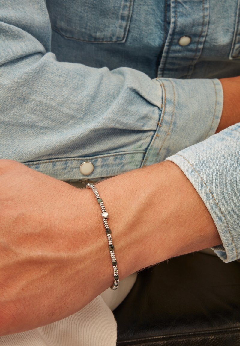 Stříbrný náramek s kulatými a srdíčkovými korálky, doplněný o zelené akcenty. Nosený na zápěstí s lehkou džínovou košilí.