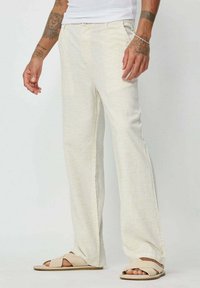Pantalons beige clair en tissu texturé, avec un design à jambe droite et un motif tissé subtil. Associés à des sandales beiges.