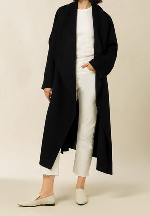 Femme portant un long manteau noir par-dessus une chemise blanche, un jean blanc court et des chaussures plates couleur crème, debout devant un fond uni.