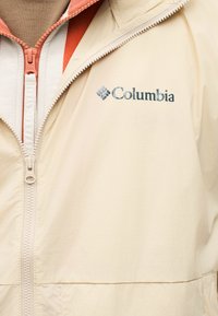 Ελαφρύ μπεζ μπουφάν Columbia με μπροστινό φερμουάρ που είναι μερικώς ανοιχτό, από κάτω στρωμένο με φλοίς σε καμένο πορτοκαλί, με ορατό φερμουάρ.