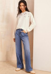 Lyse beige sweater med blomsterbroderi og lange ærmer, parret med højtaljede blå jeans. Modellen har beige slip-on sko på.