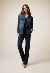 Veste en denim au design classique, haut bleu marine et pantalon bleu marine ajusté. Le mannequin porte des sandales noires à lanières et des bracelets en or.