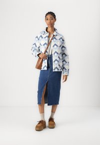 Chaqueta acolchada de patrón geométrico en azul y blanco, combinada con una falda de mezclilla con una abertura frontal. Zapatos de plataforma marrones y un bolso pequeño marrón.