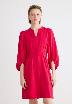 Femme portant une robe rose vif longueur genou avec des manches bouffantes froncées et un col en V, debout devant un fond blanc uni.