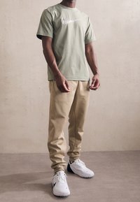 Olive zielony t-shirt z białym logo, zestawiony z beżowymi dresami i białymi butami sportowymi z czarnymi akcentami, stojący na neutralnym tle.