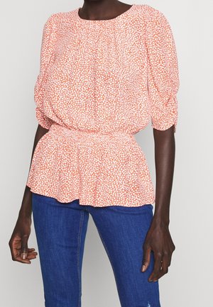 Kvinde iført en fersken- og hvidmønstret bluse med puffede ærmer og peplumtalje, kombineret med blå jeans mod en ensfarvet baggrund.