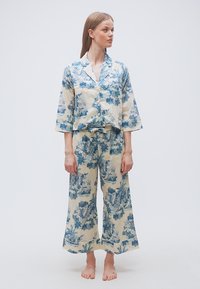 Γυναίκα που στέκεται ξυπόλυτη φορώντας κρεμ και μπλε φλοράλ φαρδιά παντελόνια και ταιριαστό πουκάμισο με κουμπιά και μανίκια τριών τετάρτων.