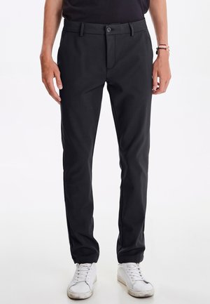 Schwarze, taillierte Hose mit geradem Schnitt, aus glattem Stoff. Verfügt über einen Knopfverschluss und seitliche Taschen. Wird mit weißen Sneakers getragen.