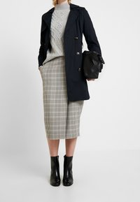 Un manteau noir sur un pull en grosse maille gris et une jupe crayon à carreaux. Bottines noires. La tenue combine des matériaux lisses et texturés.