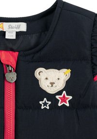 Gilet imbottito blu navy con zip rossa, caratterizzato da un patch a forma di orso e tre stelle ricamate in rosso, bianco e blu. Tessuto liscio e colletto rotondo.