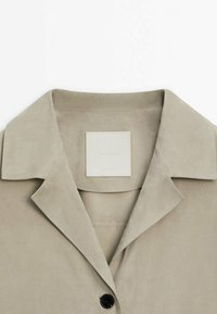 Beige nahkahame, jossa on lovettu kaulus, yksi musta nappikiinnitys ja neliön muotoinen brändilogo kauluksen sisäpuolella.