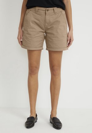 Femme portant un short beige à ourlet roulé, un haut noir et des mocassins noirs sans lacets, debout devant un fond blanc uni.
