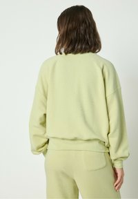 Gai zaļš lielizmēra džemperis ar raglanu piedurknēm, teksturēta auduma un ar apakšējo malžu. Valkāts ar atbilstošiem gaiši zaļiem biksēm.