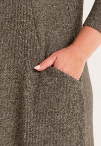 Brun ribbad klädestyp med en mjuk textur, som har en sidoficka och en synlig hand som vilar inuti.