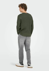 Pull en tricot torsadé vert foncé avec col rond, accompagné de jeans gris clair et de baskets blanches avec des accents noirs. Tissu texturé, coupe décontractée.