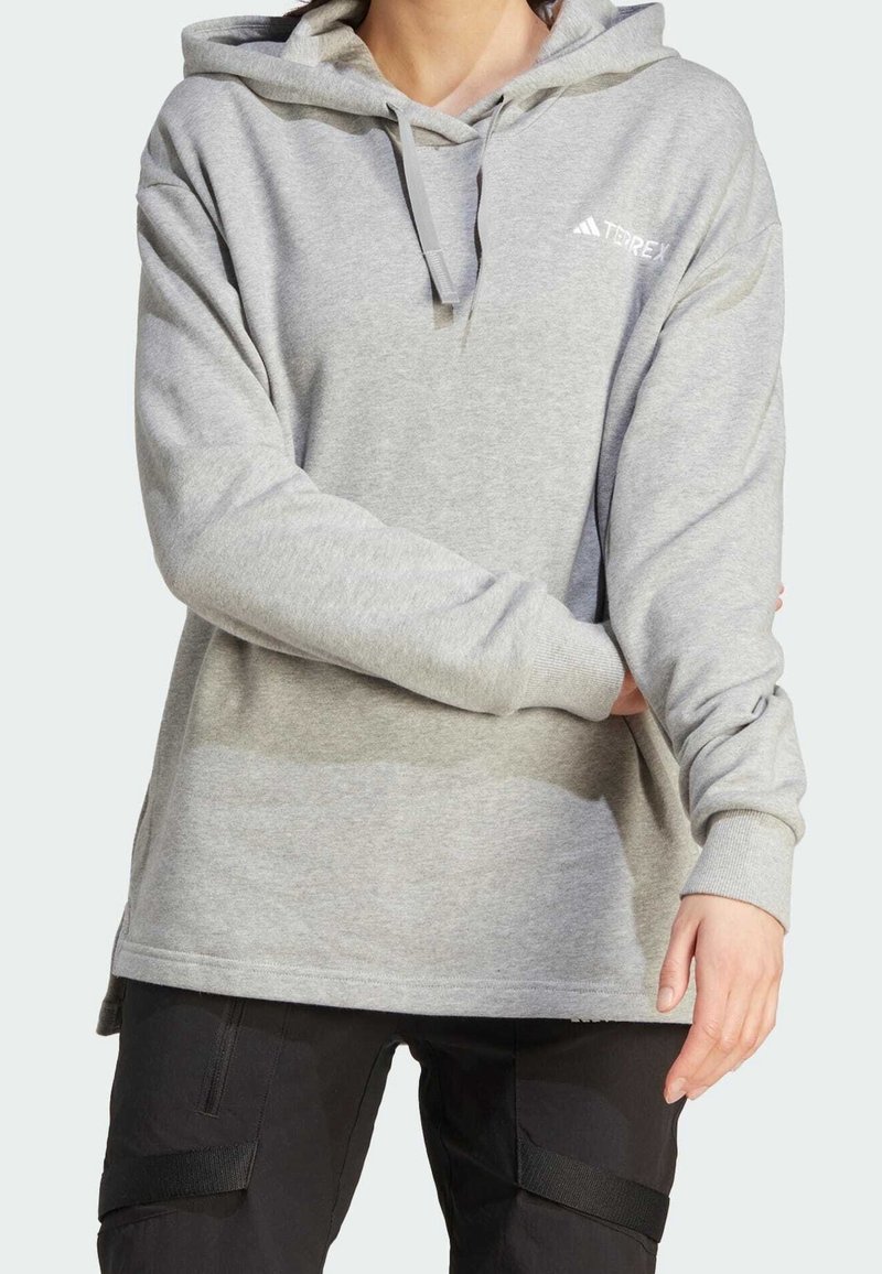 Personne portant un sweat à capuche gris clair avec le logo "TERREX" et un pantalon noir, les bras croisés à la taille, devant un fond uni.