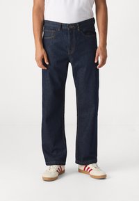 Jean en denim bleu foncé avec une coupe droite, comportant cinq poches et des coutures contrastantes, portés avec des baskets blanches rehaussées de rayures rouges.
