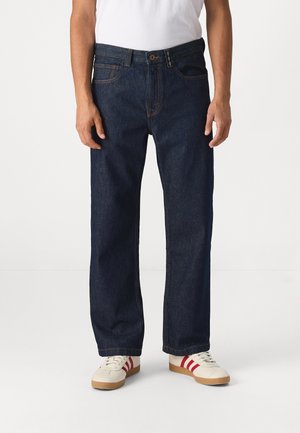 Vaqueros de mezclilla azul oscuro con un diseño de pierna recta, que cuentan con cinco bolsillos y costuras en contraste, usados con zapatillas blancas acentuadas con rayas rojas.