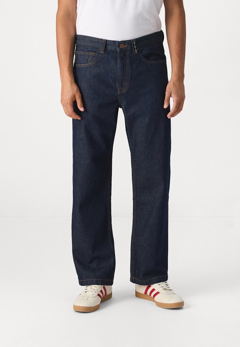 Jean en denim bleu foncé avec une coupe droite, comportant cinq poches et des coutures contrastantes, portés avec des baskets blanches rehaussées de rayures rouges.