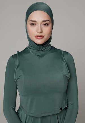 Vrouw draagt een groene aansluitende hijab en een bijpassende top met lange mouwen, kijkt recht vooruit tegen een neutrale achtergrond.