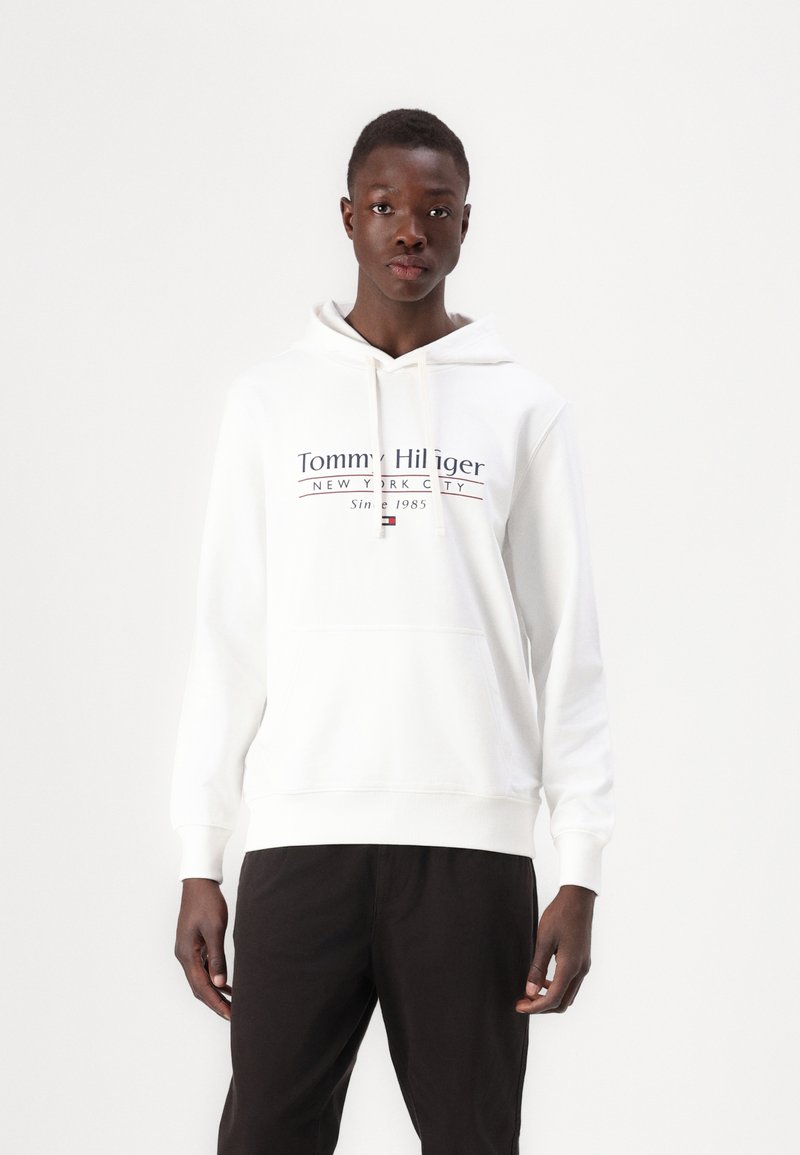 Hanorac alb cu glugă, dotat cu un buzunar frontal și șnur. Prezintă logo-ul "Tommy Hilfiger New York City" în litere bleumarin și roșii.