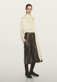 Ivory zijde top met een gedrapeerde kraag, gecombineerd met donkerbruin leren culottes en strakke bruine laarzen, gepresenteerd op een neutrale achtergrond.