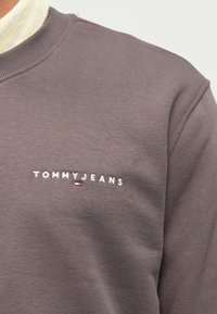 Gros plan sur un sweat-shirt marron avec le texte blanc "TOMMY JEANS" et un petit logo Tommy Hilfiger rouge, blanc et bleu sur la poitrine.