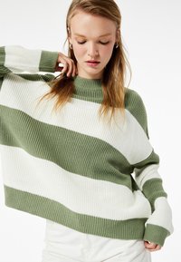 Strickpullover mit grünen und weißen horizontalen Streifen, hohem Kragen, lockerer Passform, geripptem Textur und überschnittenen Schultern.
