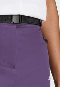 Pantaloni sportivi viola realizzati in tessuto liscio, con una cintura nera e una piccola tasca con zip di lato. Logo Adidas visibile.