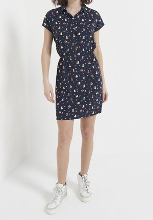 Kobieta ubrana w granatową sukienkę z krótkim rękawem z małym wzorem kolorowych kreskówkowych twarzy oraz białe wysokie trampki.