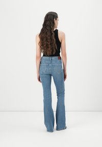 Calças de ganga boca de sino em azul claro. Apresentam uma cintura alta, design de cinco bolsos e um patch de couro castanho na parte de trás da cintura. Textura suave.