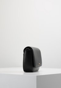 Borsa a tracolla in pelle nera testurizzata con chiusura a pattina e piccolo dettaglio dorato, esposta su un piedistallo bianco contro uno sfondo grigio chiaro.