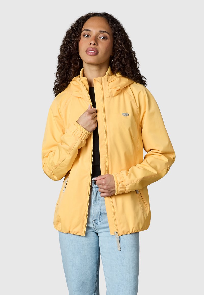 Giovane donna con capelli ricci che indossa una giacca gialla con cappuccio sopra un top nero e jeans celesti, tenendo la zip della giacca.